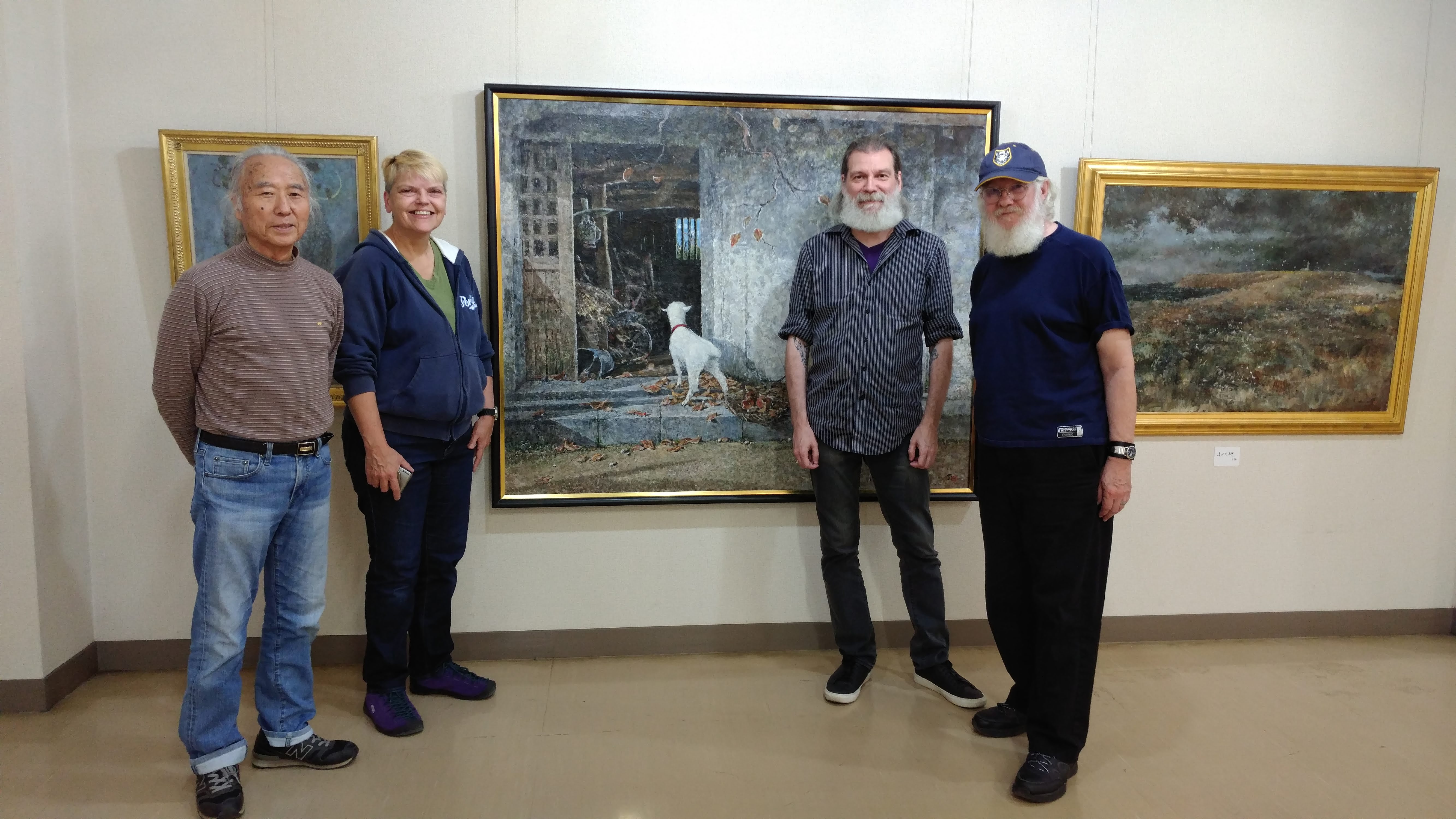 the artist, Brenda, Earl, and Chris stand in front of a painting of a goat looking into the entrance of a small stone building