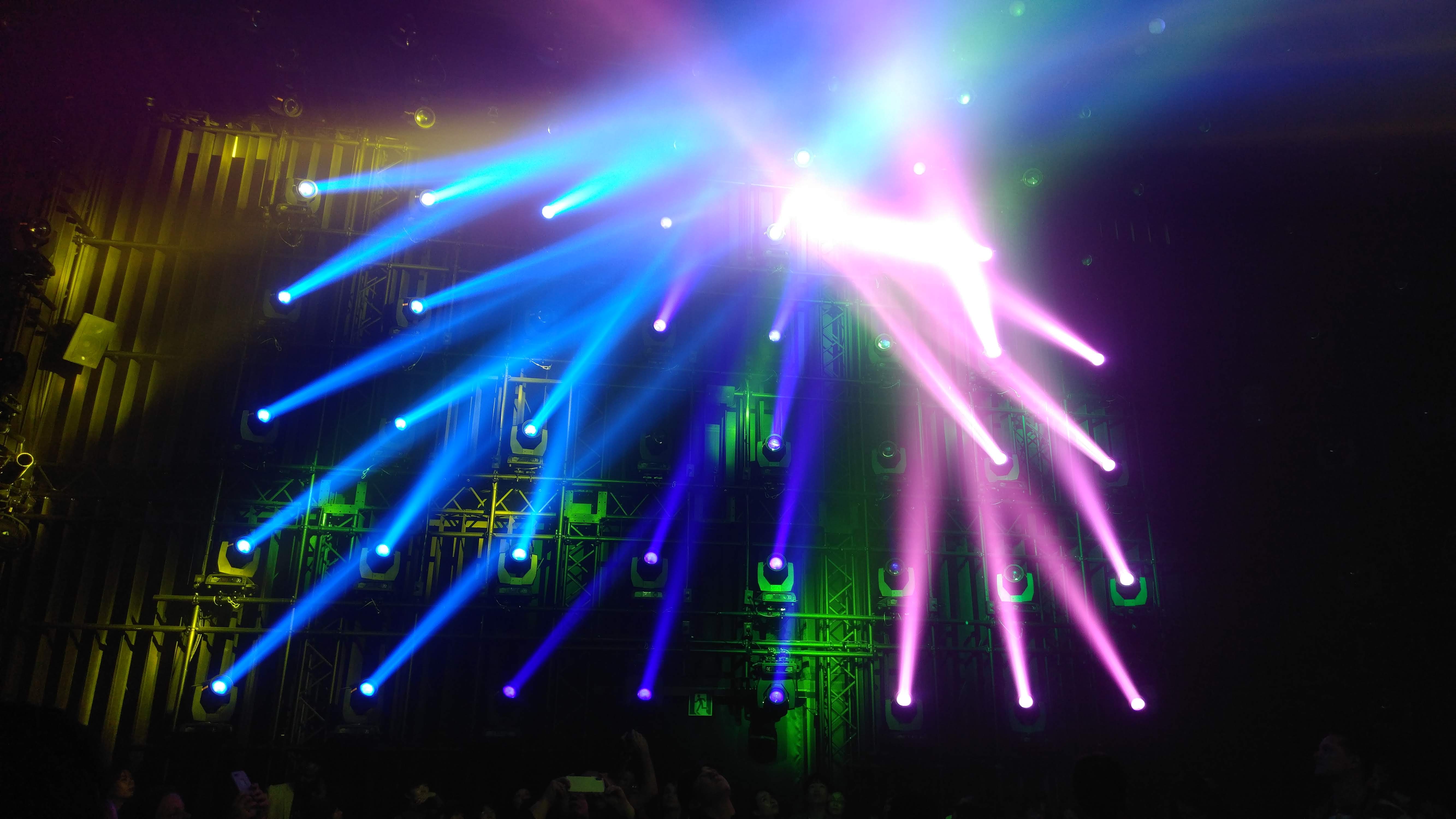 a grid of light fixtures sends pink and blue beams into a dark room.  The lighting grid is partially lit with green and yellow light.