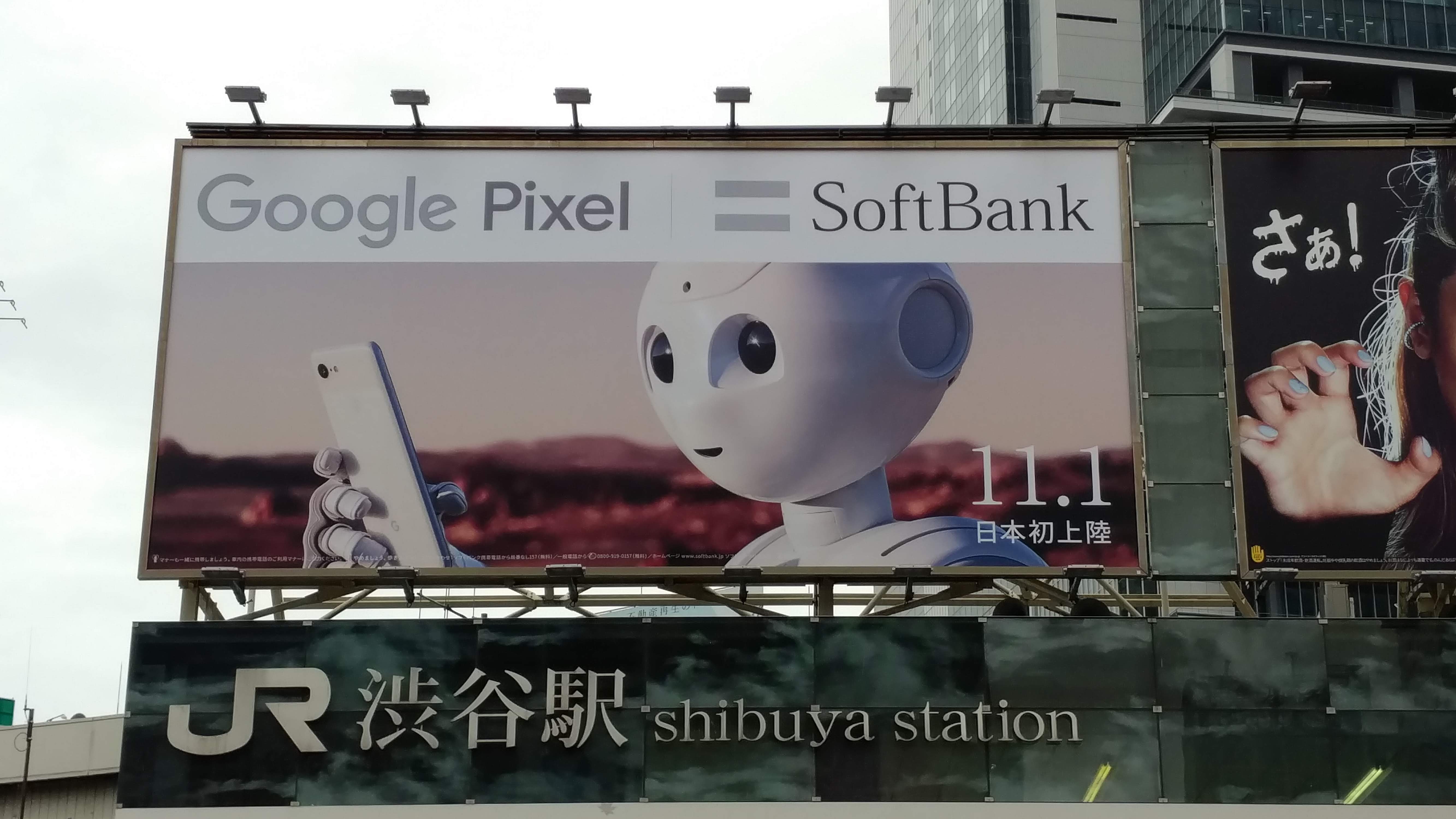 JR shibuya station billboard with text google pixel = softbank and an image of a robot holding a smartphone